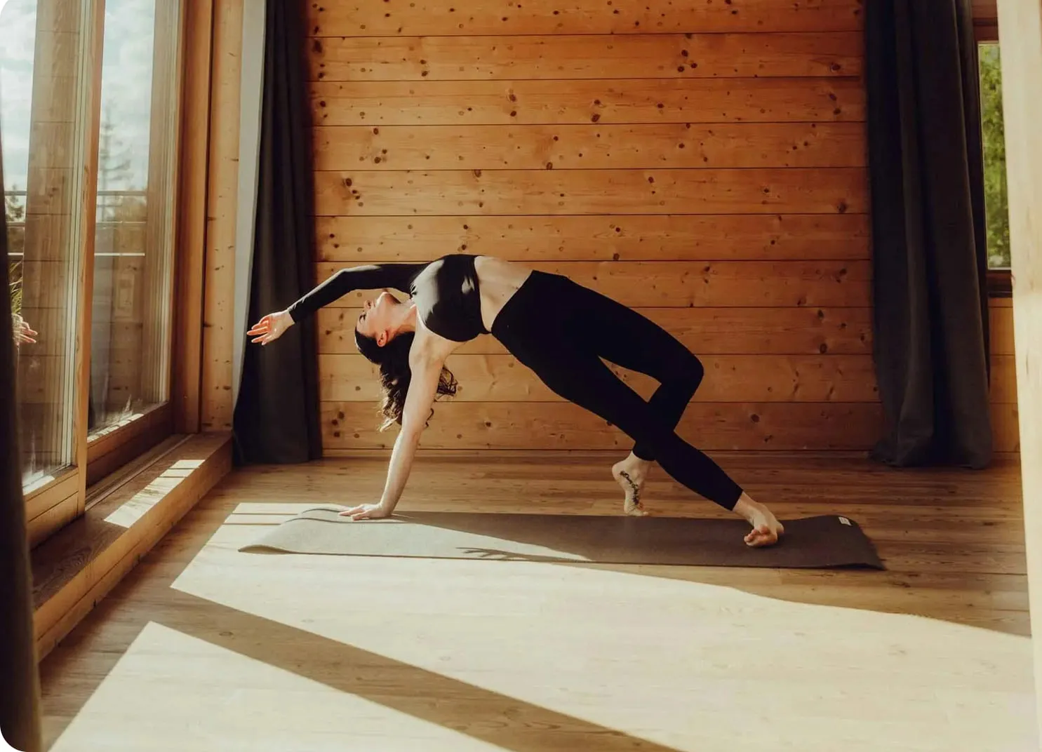 Eine Person macht Yoga auf einer Matte in einem Holzraum mit großen Fenstern.